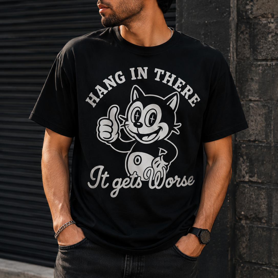 A man wearing a black graphic t-shirt with a white cartoon cat design and the text 'HANG IN THERE It gets Worse', standing against a dark textured wall.