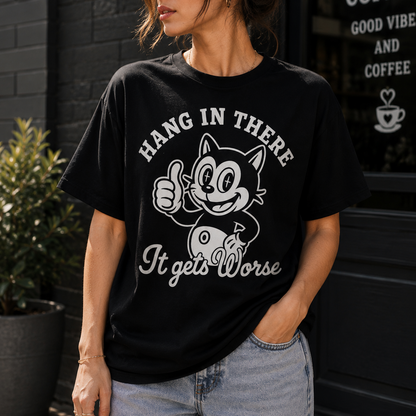A woman wearing a black graphic t-shirt with a white print of the Catwoman cartoon character and the text 'HANG IN THERE It gets Worse', paired with light wash jeans, standing in front of a dark storefront.