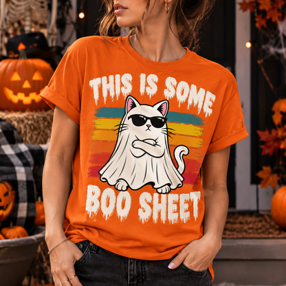 A woman wearing a bright orange graphic t-shirt with a cartoon ghost cat wearing sunglasses and the text 'THIS IS SOME BOO SHEET'. The background is a decorated Halloween porch with pumpkins, a skeleton, and autumn decorations.
