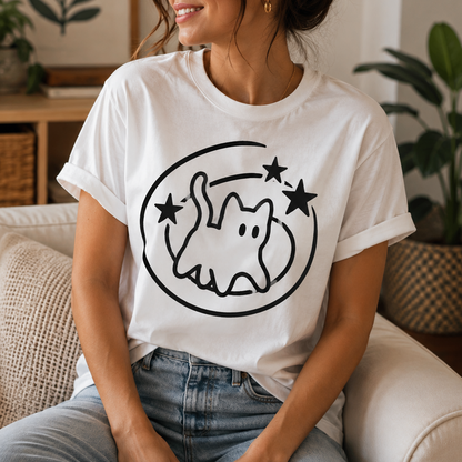A woman wearing a white graphic t-shirt with a black cat outline design, sitting on a beige couch in a cozy living room setting.
